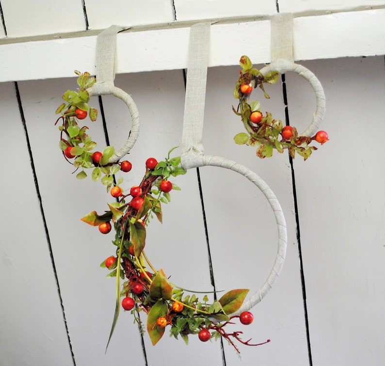 May include: Three wreaths made of woven branches with red berries and green leaves hanging from a white wall.
