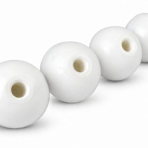 May include: Four spherical white beads arranged in a diagonal line. Each bead features a smooth, glossy finish and a centred circular hole that extends through the middle, revealing a slightly off-white interior surface within the opening.