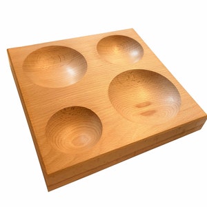 May include: A square wooden tray in a light colour, featuring four evenly spaced, rounded indentations. The tray is made of light-coloured wood, possibly beech or maple, with a smooth, finished surface. The indentations are bowl-shaped.