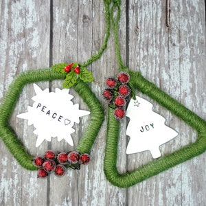 May include: Two green yarn Christmas ornaments with white snowflake and Christmas tree shapes. The ornaments have the words "Peace" and "Joy" written on them. Each ornament is decorated with red berries.