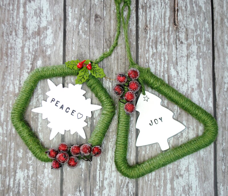 May include: Two green yarn Christmas ornaments with white snowflake and Christmas tree shapes. The ornaments have the words "Peace" and "Joy" written on them. Each ornament is decorated with red berries.