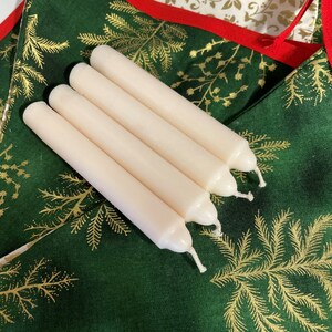 Peut inclure: Quatre bougies cylindriques blanc cass&eacute; avec des sommets arrondis et des m&egrave;ches en coton visibles. Les bougies sont dispos&eacute;es en rang&eacute;e parall&egrave;le, chacune pr&eacute;sentant une surface en cire lisse et solide. Elles sont pos&eacute;es sur un tissu vert fonc&eacute;.