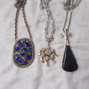 May include: Three necklaces with silver chains. One has an oval pendant with blue gemstones. Another features a flower-shaped pendant with iridescent crystals. The third has a black, triangular pendant.