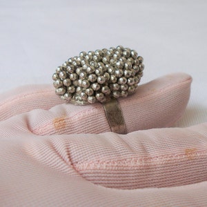 May include: A silver ring featuring a cluster of small, round beads. The ring's band is a simple, thin silver band. The beaded cluster is the focal point, creating a unique and textured appearance. The ring is shown on a pink surface.