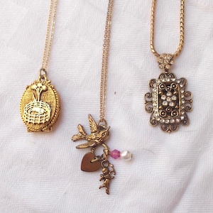 May include: Three gold-toned necklaces with different pendants. One has an oval pendant with a cage design, another features a bird with a heart and cherub, and the third has a rectangular pendant with rhinestones.