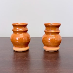 May include: Two small, handmade ceramic vases with a warm, reddish-brown glaze. Each vase has a rounded body, a narrow neck, and a flared rim. The vases sit on a light brown base, and are displayed on a dark brown surface.