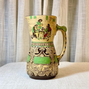 May include: Vintage ceramic pitcher with a cream-colored base, featuring a detailed scene of figures at a table. The pitcher has a green handle and accents, with ornate floral and scrollwork designs in brown and gold. The rim is dark brown.