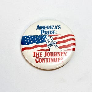 May include: A round button with the text "America's Pride: The Journey Continues" in blue and red. The button features a stylized American flag and a space shuttle graphic.