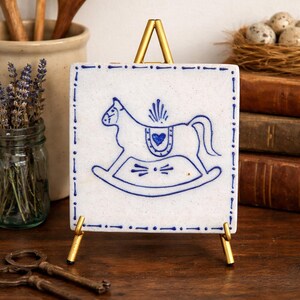 May include: A square, white ceramic tile with a blue rocking horse design, displayed on a small gold easel. The tile has a blue border and is surrounded by other decorative items, including old books and keys.