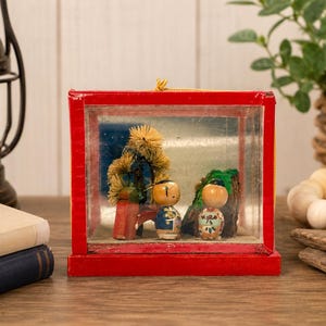 May include: A rectangular red wooden display case with a clear front panel containing two small wooden figurines, a miniature tree, and a rock formation. The figurines feature painted details and are positioned on a light-colored base inside the frame.