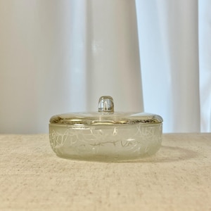 May include: A vintage glass trinket box with a silver-toned rim and a clear, rectangular handle on the lid. The frosted glass features a delicate, white, swirling pattern. The box is oval-shaped and sits on a neutral surface.