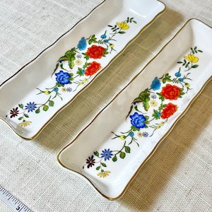 May include: Two rectangular, white ceramic serving dishes with a floral design. The floral pattern includes red, blue, and yellow flowers with green leaves. The edges are trimmed with gold. The dishes are approximately 8 inches long.