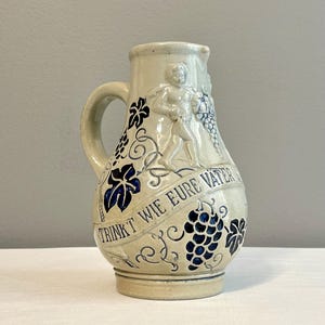May include: A beige ceramic pitcher with a handle, featuring blue grape and leaf designs. The pitcher has a relief of a man and the text "TRINKT WIE EURE VATER". A classic piece for serving beverages.