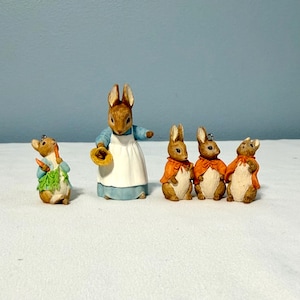 May include: A set of five rabbit figurines. One rabbit wears a blue jacket and holds carrots. Another is a larger rabbit in a blue dress and white apron holding a straw hat. Three smaller rabbits wear orange capes and have light-colored bellies.