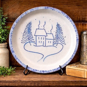 May include: A white ceramic plate with a blue rim and a hand-painted winter scene. The design features two houses, two pine trees, and a winding path, all in blue. The plate is displayed on a black metal stand.