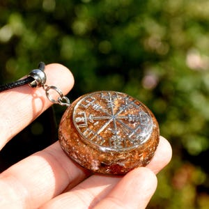 Op de afbeelding: Een ronde hanger met een zilverkleurig Vegvisir-symbool ingebed in een getextureerde, barnsteenkleurige harsbasis. De hanger is bevestigd aan een zwart koord met een zilverkleurige metalen hangerhouder, wat een cirkelvormig halssieraad vormt.