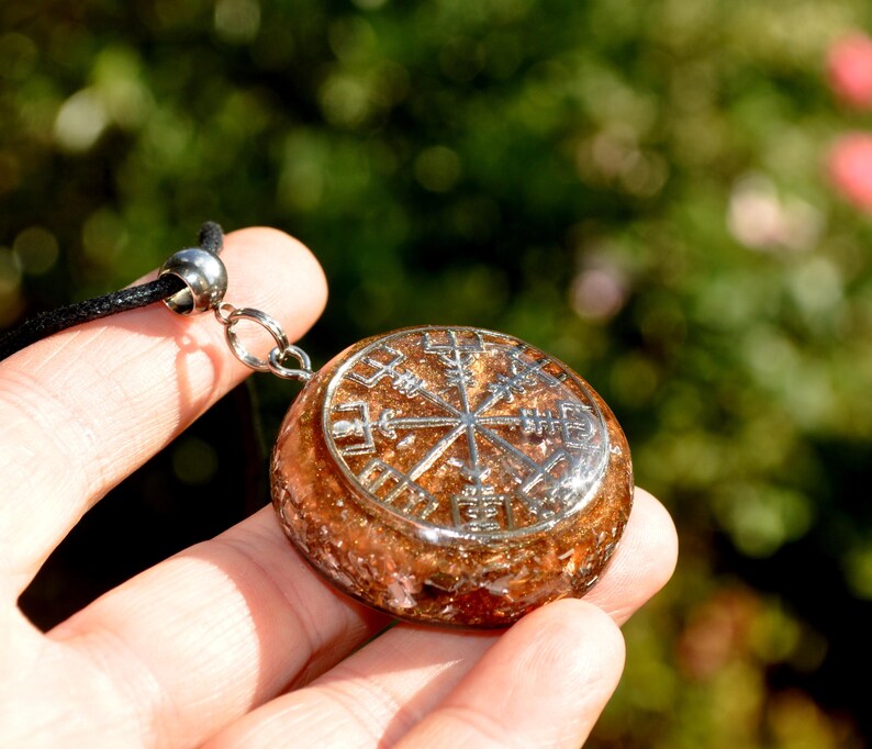 Op de afbeelding: Een ronde hanger met een zilverkleurig Vegvisir-symbool ingebed in een getextureerde, barnsteenkleurige harsbasis. De hanger is bevestigd aan een zwart koord met een zilverkleurige metalen hangerhouder, wat een cirkelvormig halssieraad vormt.