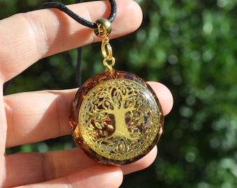 May include: A round pendant featuring a gold-colored tree of life emblem embedded within a translucent amber-toned resin base. The pendant is attached to a black cord by a gold-colored metal bail and jump ring connector.