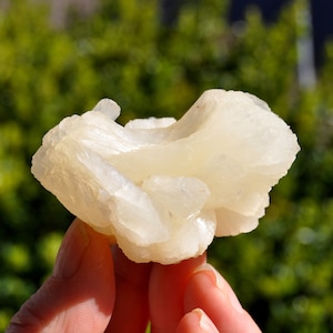 Könnte beinhalten: Ein rohes, durchscheinendes weißes Mineralexemplar mit einer Ansammlung kristalliner Formationen. Der Stein weist eine geschichtete, fächerartige Struktur mit glatten, reflektierenden Oberflächen und unregelmäßigen Kanten auf.