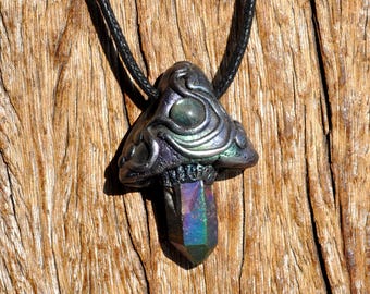 May include: A pendant shaped like a mushroom with a dark, iridescent cap featuring swirling raised patterns and a central circular accent. A faceted, multi-colored crystal point hangs from the base of the cap, suspended by a black cord.