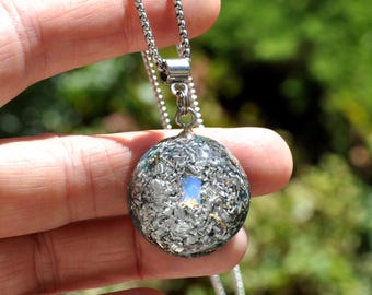 May include: A circular pendant necklace featuring a clear resin dome filled with metallic silver shavings and a single iridescent opal-like stone at the center, suspended from a silver-toned metal box chain with a polished finish.