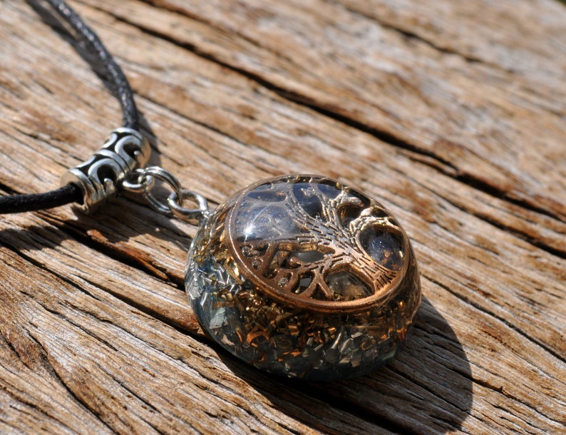 Puede incluir: Un colgante redondo de cristal transparente con un dise&ntilde;o de &aacute;rbol de la vida dorado. El colgante est&aacute; en un cord&oacute;n de cuero negro con un cierre plateado.