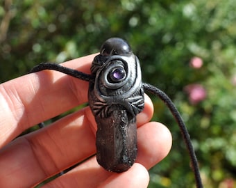 May include: A dark, raw crystal pendant featuring a silver-toned metallic wrap with leaf-like textures and a small, round, purple gemstone set in a spiral design. The pendant is suspended from a thin black cord, showcasing a natural, jagged base.