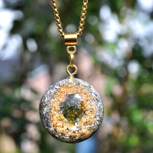Op de afbeelding: Een ronde hanger met een donkergroen steenfragment in het midden, omringd door bladgoudvlokken en zilveren metaalsplinters, ingebed in een heldere harsbol. De hanger hangt aan een goudkleurige ketting met een cilindervormig oogje.
