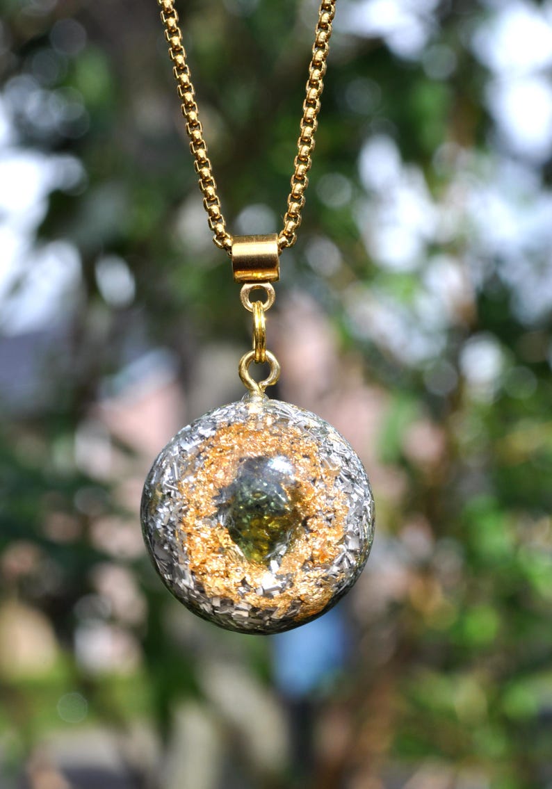 Op de afbeelding: Een ronde hanger met een donkergroen steenfragment in het midden, omringd door bladgoudvlokken en zilveren metaalsplinters, ingebed in een heldere harsbol. De hanger hangt aan een goudkleurige ketting met een cilindervormig oogje.