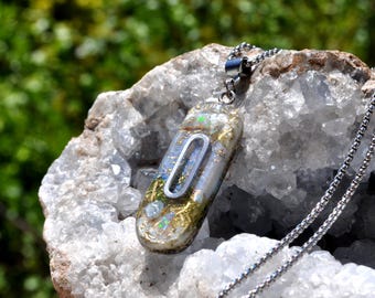 May include: A pendant necklace with a rectangular, clear resin pendant. The pendant has a silver-colored frame with a blue and gold design inside. The necklace chain is silver-colored and rests on a white and gray crystal.