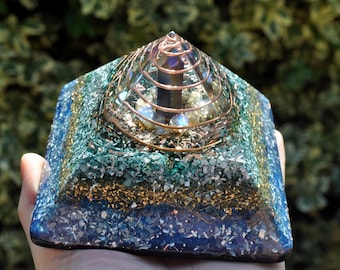 May include: A pyramid-shaped resin object featuring layers of blue, green, and gold metallic flakes. A copper wire coil spirals around a central dark crystal point embedded at the apex, surrounded by small, light-colored spherical stones.