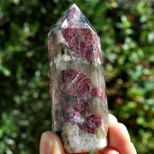 May include: A polished crystal tower with a pointed top. The crystal is a mix of clear, gray, and white, with large, irregular patches of deep red. The crystal is held in a hand against a blurred green background.