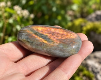 May include: A smooth, oval-shaped labradorite gemstone featuring a polished surface with a prominent iridescent flash of orange and gold light across its gray, multi-toned base. The stone displays natural internal striations and color variations.