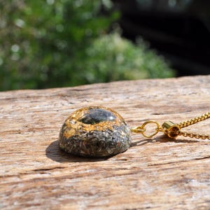 Op de afbeelding: Een koepelvormige hanger aan een ketting met een donkere, getextureerde basis met ingebedde metaalvlokken en een donker steenaccent in het midden, hangend aan een goudkleurige ketting. De hanger heeft een glad, afgerond oppervlak en een gouden oogje.