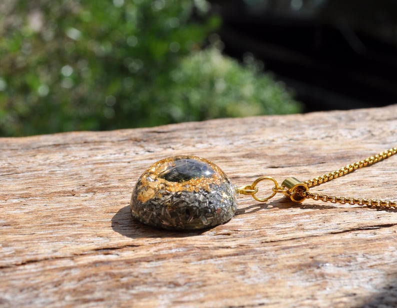 Op de afbeelding: Een koepelvormige hanger aan een ketting met een donkere, getextureerde basis met ingebedde metaalvlokken en een donker steenaccent in het midden, hangend aan een goudkleurige ketting. De hanger heeft een glad, afgerond oppervlak en een gouden oogje.