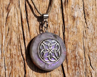 May include: A round pendant necklace featuring a silver-toned metal Celtic knot emblem embedded in a translucent, marbled purple and grey resin disc. The pendant hangs from a thin, light brown cord with a silver-toned metal bail connector.