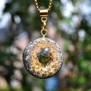 Op de afbeelding: Een bolvormige hanger met een donkergroen steenfragment in het midden, omringd door bladgoud en zilverkleurige metaalsnippers, ingesloten in een heldere harsbol en hangend aan een goudkleurige metalen ketting.