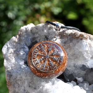 Op de afbeelding: Een ronde hanger met een zilverkleurig Vegvisir-symbool ingebed in een getextureerde, barnsteenkleurige harsbasis. De hanger is bevestigd aan een zwart koord met een zilverkleurige metalen hangerhouder, wat een cirkelvormig sieraad vormt.