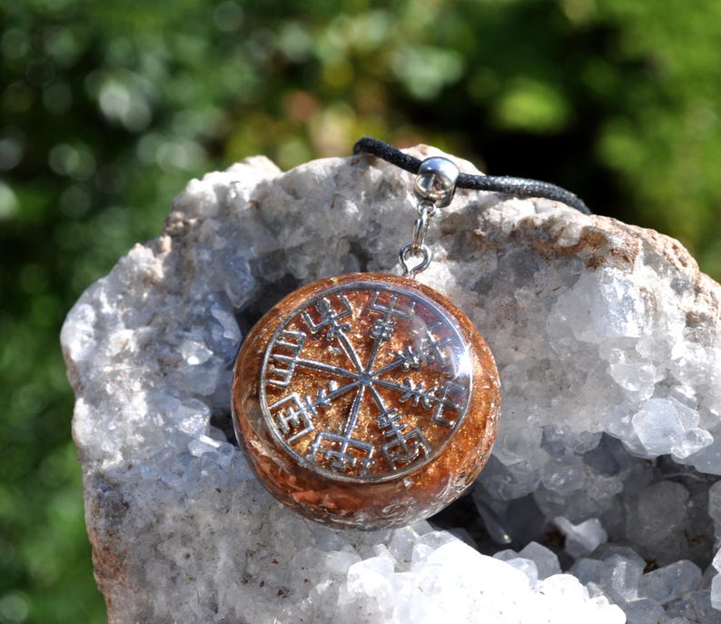 Op de afbeelding: Een ronde hanger met een zilverkleurig Vegvisir-symbool ingebed in een getextureerde, barnsteenkleurige harsbasis. De hanger is bevestigd aan een zwart koord met een zilverkleurige metalen hangerhouder, wat een cirkelvormig sieraad vormt.