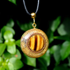 May include: A circular pendant featuring a central tiger's eye gemstone with brown and gold bands, encircled by a twisted copper wire. The stone is embedded in a translucent, textured resin disc with gold-toned flecks, suspended from a black cord.