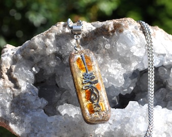 May include: A rectangular pendant necklace featuring a clear resin body embedded with amber-colored fragments and a silver-toned metal unalome symbol. The pendant hangs from a silver-toned metal chain, showcasing a polished finish and intricate internal details.