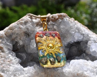 May include: A rectangular pendant featuring a gold-toned sun emblem at the center, surrounded by embedded metallic-colored stones. The pendant has a gradient background transitioning from red at the top to blue at the bottom, suspended by a cord.