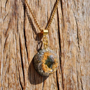 Op de afbeelding: Een ronde hangerketting met een goudkleurige ketting en een harsbedel. De bedel bevat ingebedde metallic zilveren vlokken, bladgoudaccenten en een centrale donkergroene gefacetteerde steen, vastgezet met een goudkleurige metalen hangerhouder.