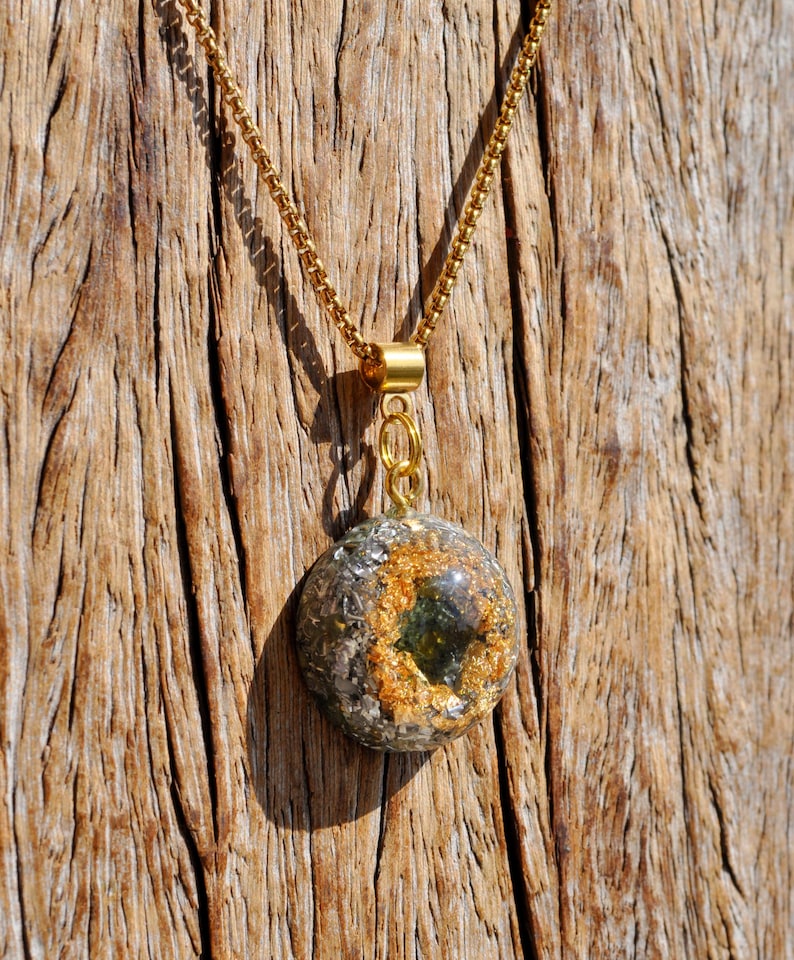 Op de afbeelding: Een ronde hangerketting met een goudkleurige ketting en een harsbedel. De bedel bevat ingebedde metallic zilveren vlokken, bladgoudaccenten en een centrale donkergroene gefacetteerde steen, vastgezet met een goudkleurige metalen hangerhouder.