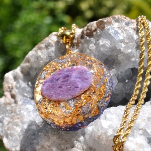 May include: A gold chain necklace with a pendant. The pendant is round, featuring a central purple stone, encircled by gold and silver flakes. The pendant rests on a white and grey rock.