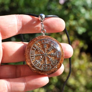 Op de afbeelding: Een ronde, barnsteenkleurige hars hanger met een ingebed zilverkleurig Vegvisir-symbool. De hanger is bevestigd aan een eenvoudige zilverkleurige metalen hangerhouder en hangt aan een dun zwart koord met een textuurrijke, glinsterende afwerking.
