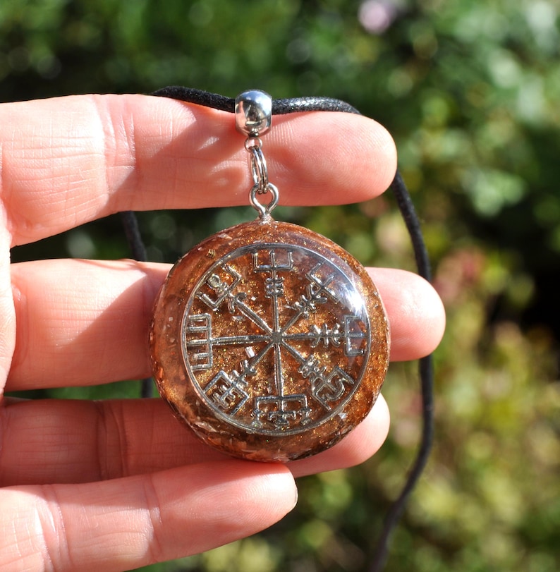 Op de afbeelding: Een ronde, barnsteenkleurige hars hanger met een ingebed zilverkleurig Vegvisir-symbool. De hanger is bevestigd aan een eenvoudige zilverkleurige metalen hangerhouder en hangt aan een dun zwart koord met een textuurrijke, glinsterende afwerking.