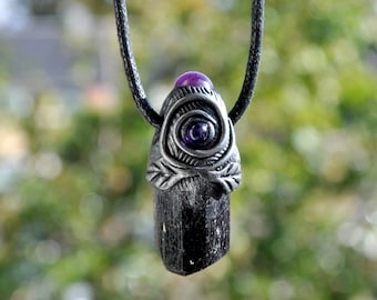 May include: A black tourmaline crystal pendant suspended on a black cord. The top of the crystal is encased in a silver-toned metal cap featuring a spiral design, a small purple bead at the peak, and a central purple stone inlay.