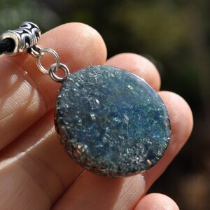 Puede incluir: Un colgante redondo de plata con una incrustaci&oacute;n de resina azul oscuro y verde. La resina tiene un efecto brillante y brillante.