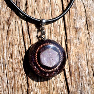 May include: A round pendant necklace featuring a dark purple resin base with embedded metallic flecks. A hexagonal light purple crystal sits in the centre, encircled by a twisted copper wire coil. The pendant hangs from a smooth black cord.
